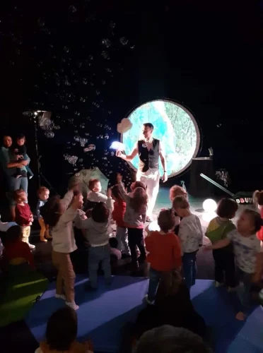 Magie Sam sur scène devant un groupe d’enfants, avec un décor circulaire lumineux.